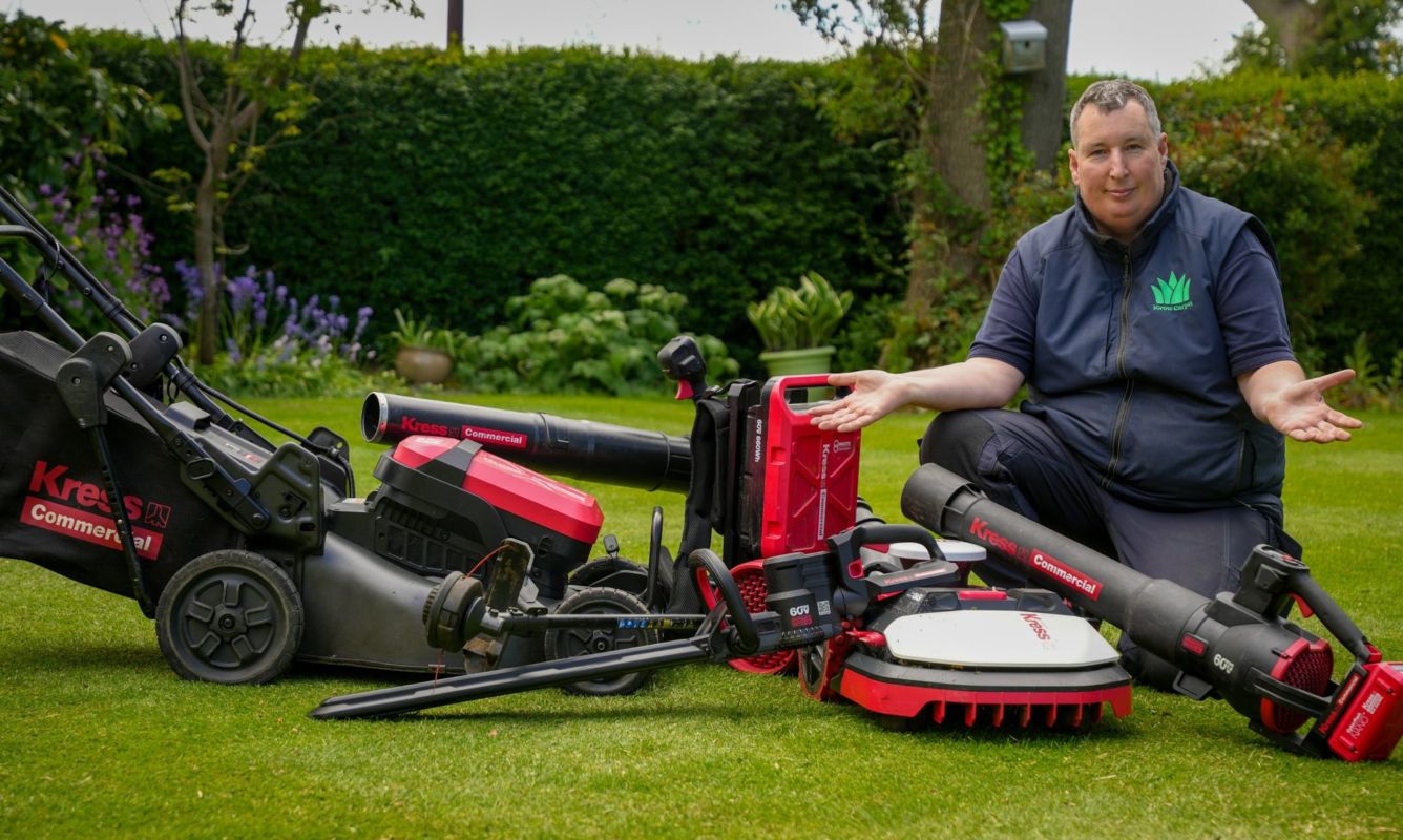 Robbie Lynn kneeling on a lawn beside a line-up of Kress Commercial battery tools, including a mower and robot mower
